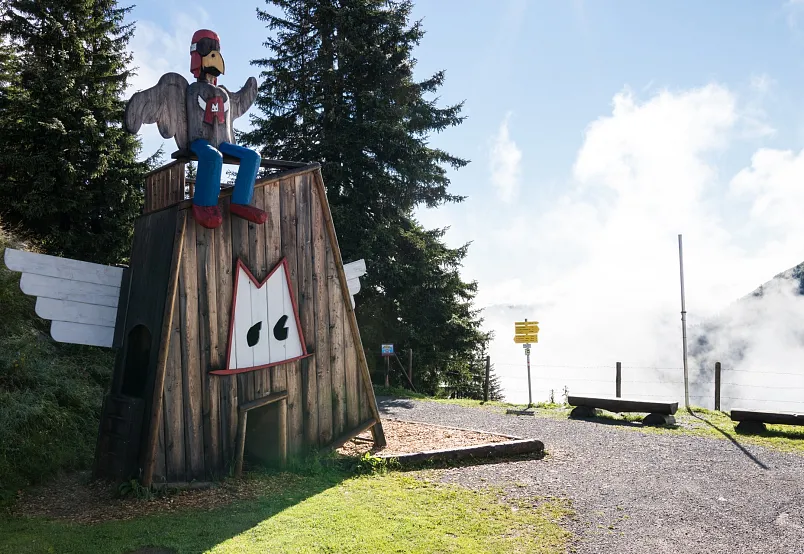 Holzskulptur eines Hahns auf einem Holzhäuschen im Grünen am Wanderweg in den Alpen.