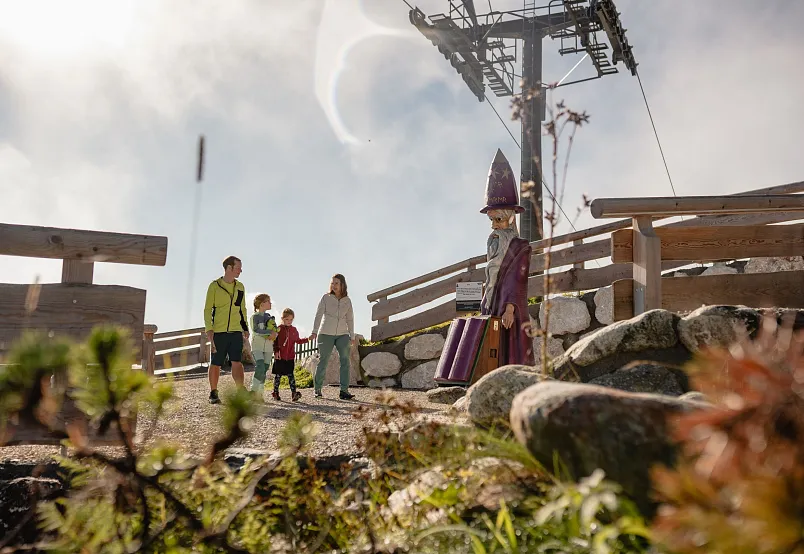 Familie wandert auf einem Bergpfad in den Alpen bei einer Holzfigur, im Hintergrund eine Seilbahn.