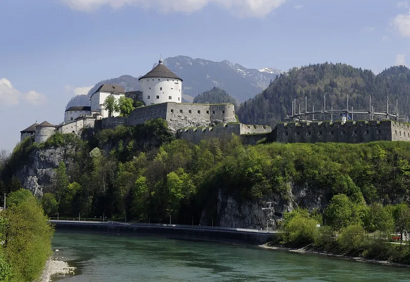 Medieval castle on a hill with mountains in the background and a river in the foreground.