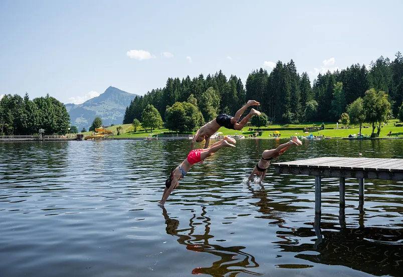 Drei Personen springen von einem Steg in einen See, umgeben von Wäldern und Bergen bei klarem Himmel.