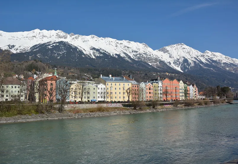 Bunte Häuserreihe am Ufer eines Flusses vor schneebedeckten Alpen in Innsbruck.