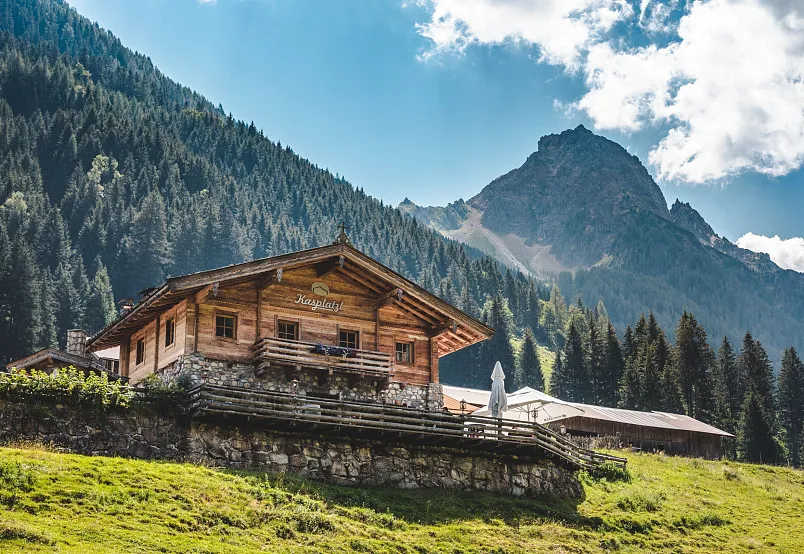 A wooden mountain cabin on a grassy hillside with trees and a rocky peak in the background under a partly cloudy sky.