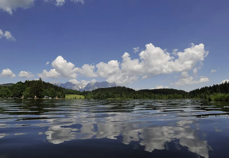 Ein ruhiger See mit sonnigem Himmel, umgeben von grünen Hügeln und bewaldeten Bergen im Hintergrund.