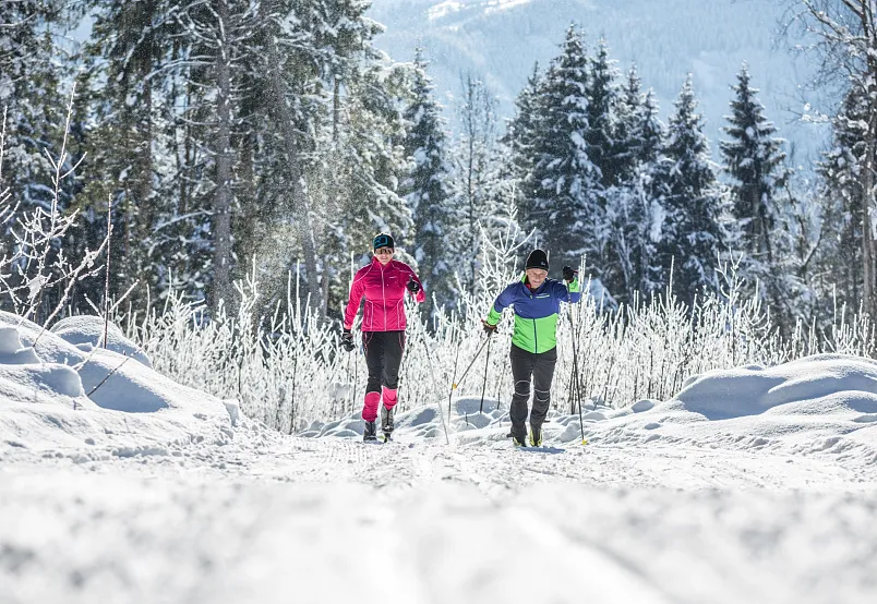 Zwei Personen beim Skilanglauf in einer verschneiten, bewaldeten Alpenlandschaft.