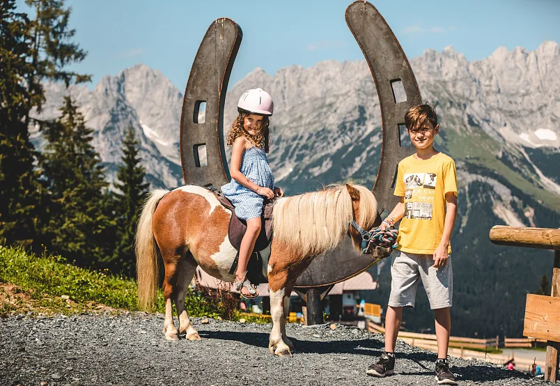 Ein Mädchen auf einem Pony und ein Junge in den Alpen mit Hufeisen im Hintergrund.