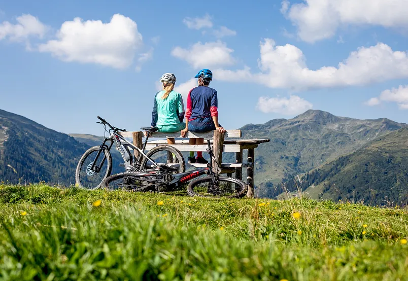 Zwei Radfahrer sitzen auf einer Bank mit Blick auf die Kitzbüheler Alpen und ihre Fahrräder neben sich.