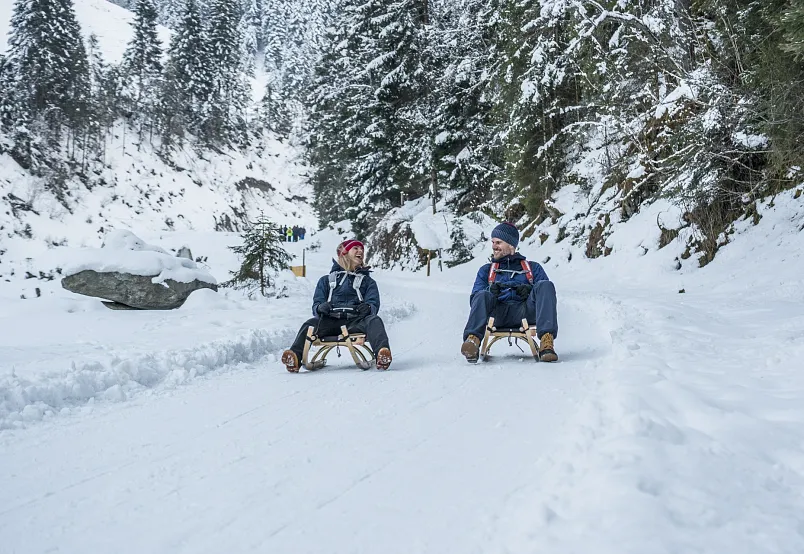 Zwei Personen rodeln auf einem verschneiten Waldweg in den Alpen.