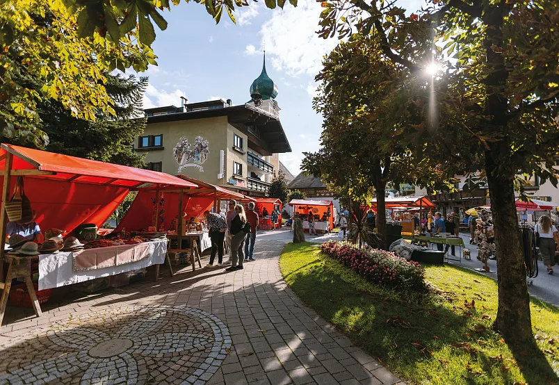 A bustling outdoor market in a picturesque town with red stalls and people walking, surrounded by trees and traditional architecture.