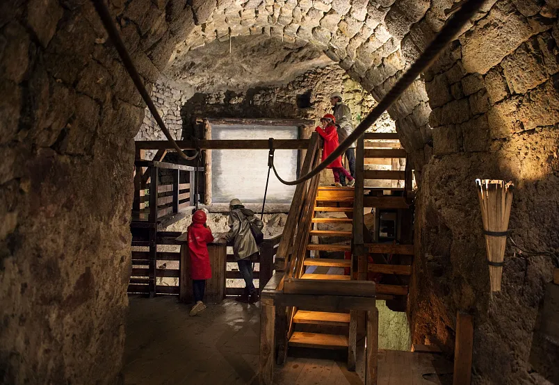 Three people in medieval clothing standing inside an underground stone chamber with wooden stairs.