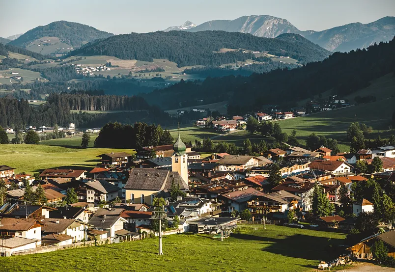 Luftaufnahme eines Dorfes in einer Berglandschaft mit grünen Wiesen und umliegenden Alpen.