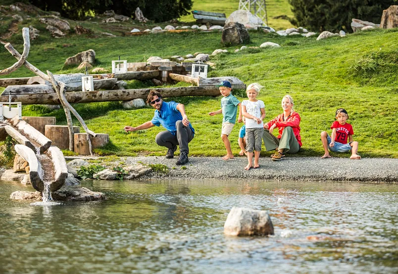 Familie mit Kindern spielt an einem flachen Teich in einer bergigen Region.