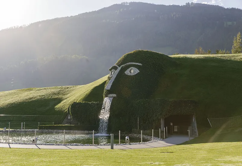 A giant grass-covered head sculpture with water flowing from its mouth in front of a mountainous landscape.