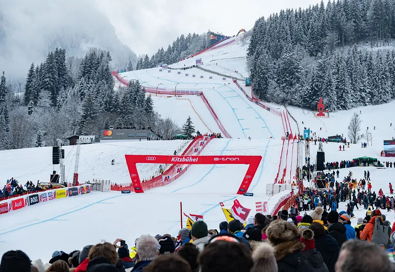Ski race event in a snowy alpine setting with spectators, a red finish line arch, and forested mountains in the background.