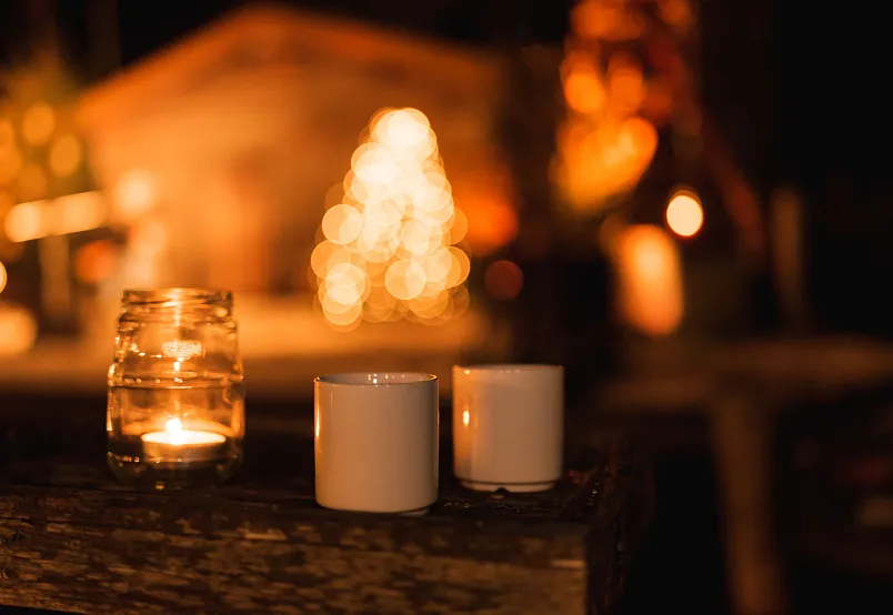Two white mugs and a candle jar on a wooden table, with blurred festive lights in the background.
