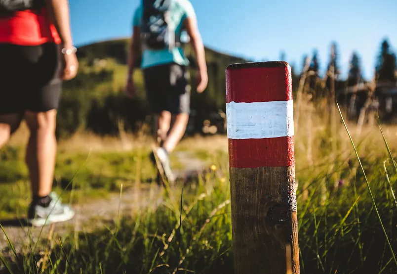Zwei Personen wandern auf einem sonnigen Pfad in einer alpinen Landschaft, im Vordergrund ein rot-weiß markierter Wanderpfosten.