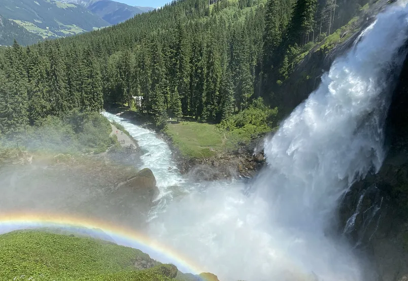 Wasserfall in alpiner Landschaft mit Regenbogen und dichten Nadelwäldern im Hintergrund.