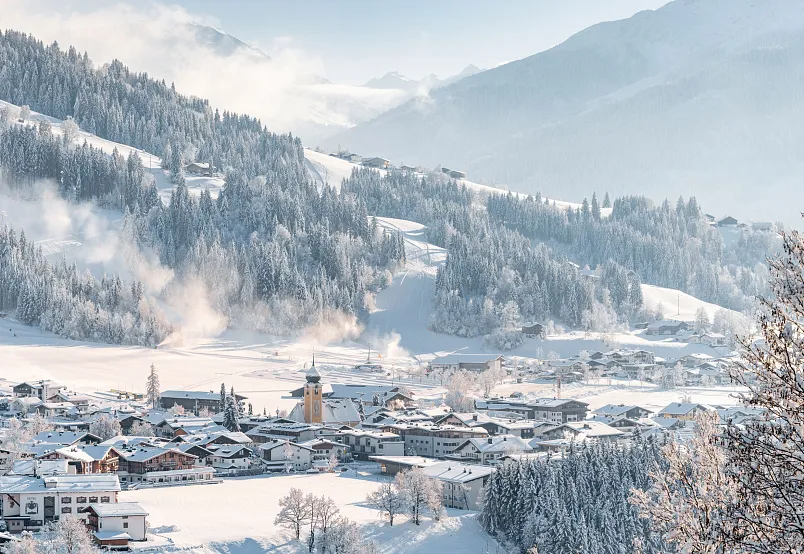 Verschneites Dorf in einer Alpenregion, umgeben von Bergen und schneebedeckten Wäldern.
