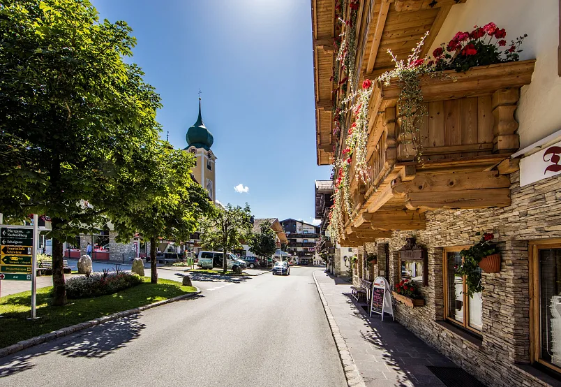 Straße in Kitzbühel mit traditionellen Gebäuden, Blumen und Kirchturmsicht an einem sonnigen Tag.