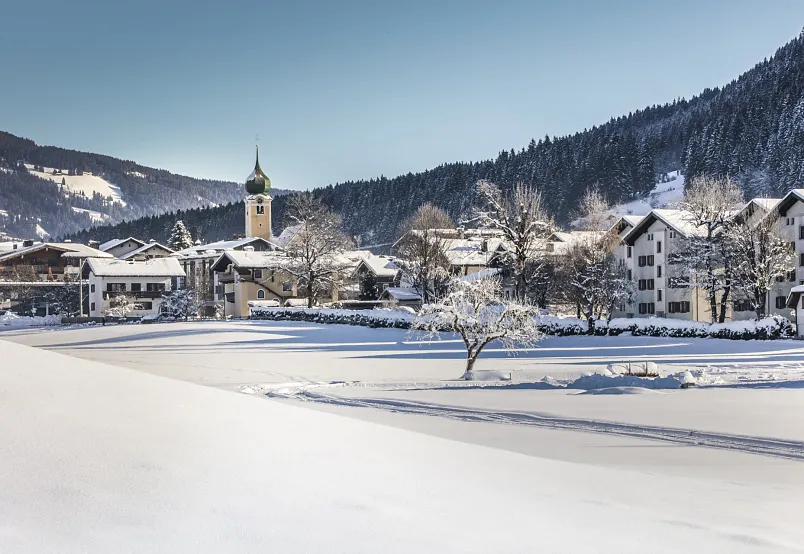 Verschneite Berglandschaft mit Dorfkirche in den Alpen, umgeben von Wäldern und Hängen, unter klarem Himmel.