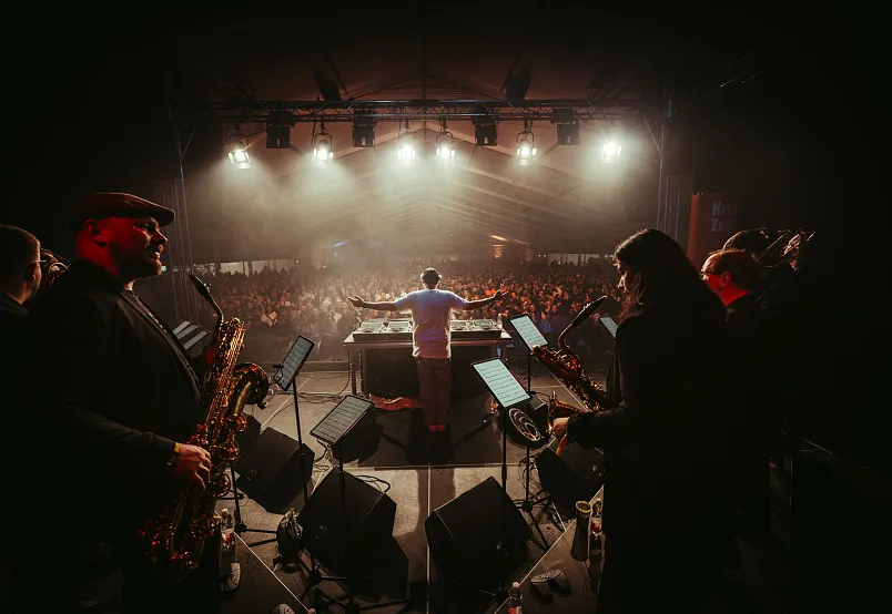 A live music performance on stage with a band playing instruments and a crowd in the background.