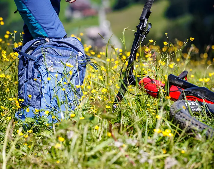 A mountain bike rests on a field of yellow wildflowers with a nearby blue backpack, suggesting a pause during a biking trip.
