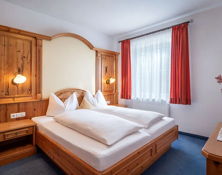 Gemütliches Hotelzimmer mit Doppelbett, Holzverkleidung und großen Fenstern mit roten Vorhängen.