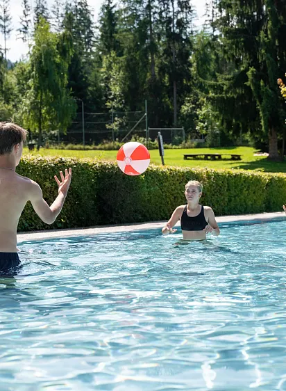 Drei junge Menschen spielen mit einem Ball im Außenpool, umgeben von Bäumen und Rasen.