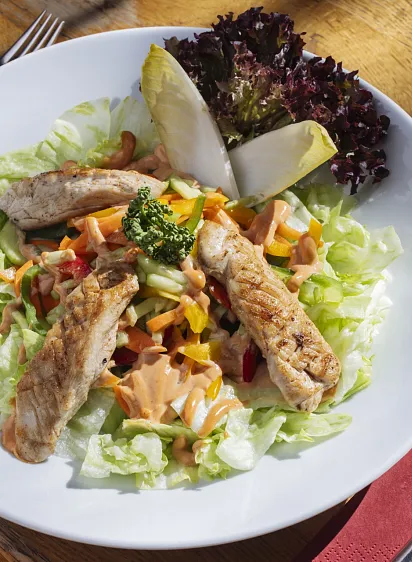 Ein Teller mit gegrilltem Hähnchen auf frischem Salat, garniert mit Gemüse und Salatblättern, auf einem hölzernen Tisch serviert.