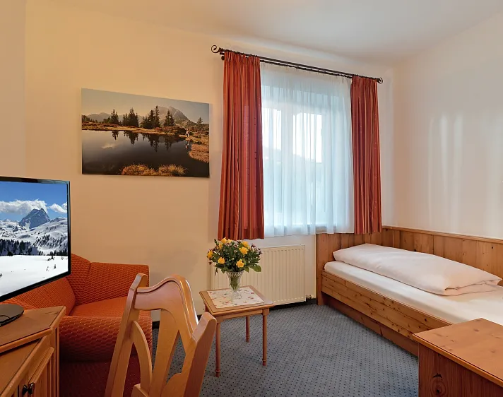 Een hotelkamer met een eenpersoonsbed, stoel, tafel, bloemen en een tv met berglandschap.