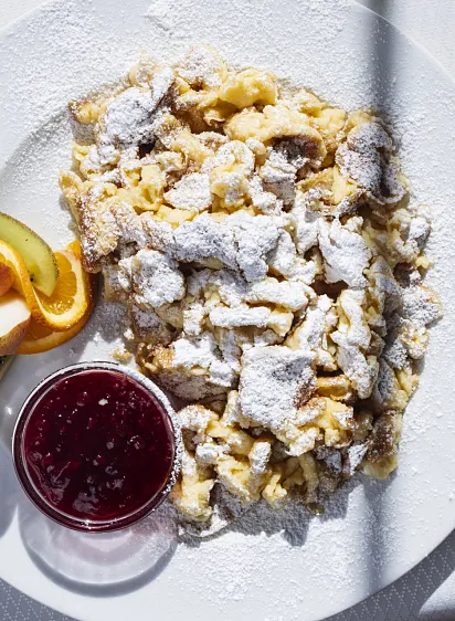 Kaiserschmarrn mit Puderzucker, Orangenscheiben, Kiwi und Marmelade auf einem Teller.