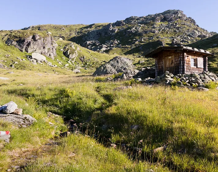 Eine Holzhütte in einer alpinen Berglandschaft mit grasbewachsenen Hügeln und einem klaren Himmel.