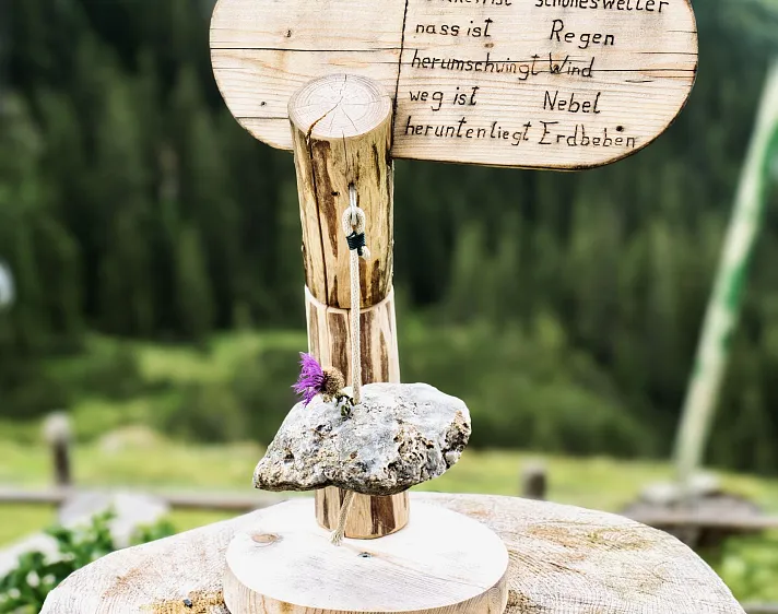 Holzschild auf einem Berg mit handgeschriebener Wettervorhersage und dekorativem Stein im Vordergrund.