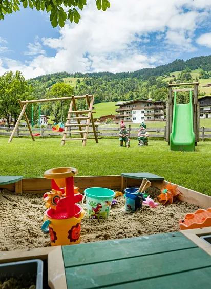 Ein Sandkasten im Vordergrund mit Spielzeug, im Hintergrund eine Schaukel, Rutsche und Alpenlandschaft in Tirol.