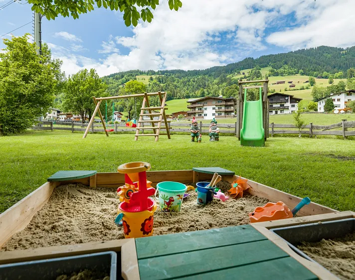 Ein Sandkasten im Vordergrund mit Spielzeug, im Hintergrund eine Schaukel, Rutsche und Alpenlandschaft in Tirol.