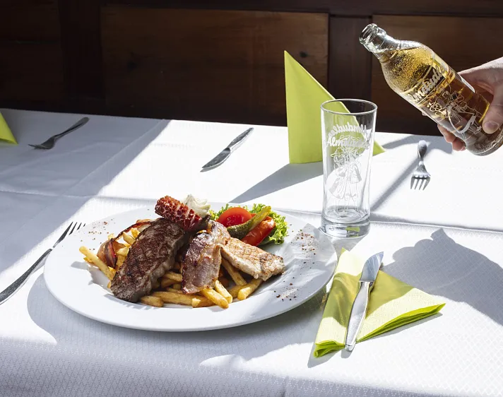 Grillfleisch und Pommes Frites auf einem Teller mit einem Glas Almdudler auf einem weißen Tisch.