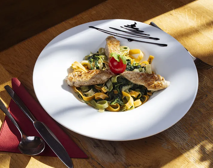 Tagliatelle mit Hähnchenstreifen, Spinat und einer Tomate auf einem weißen Teller.