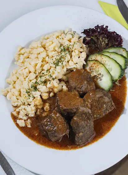 Teller mit Rindsgulasch, Spätzle, Gurkenscheiben und Salat.