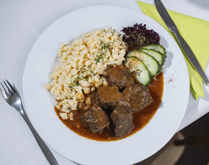 Teller mit Rindsgulasch, Spätzle, Gurkenscheiben und Salat.