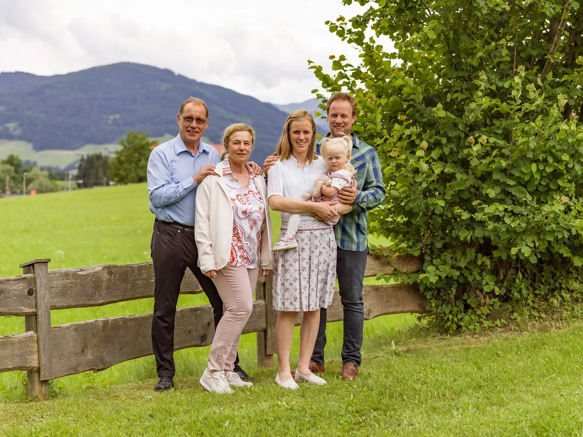 Fünf Personen stehen auf einer Wiese vor einem Holzzaun in einer ländlichen Umgebung mit Hügeln im Hintergrund.