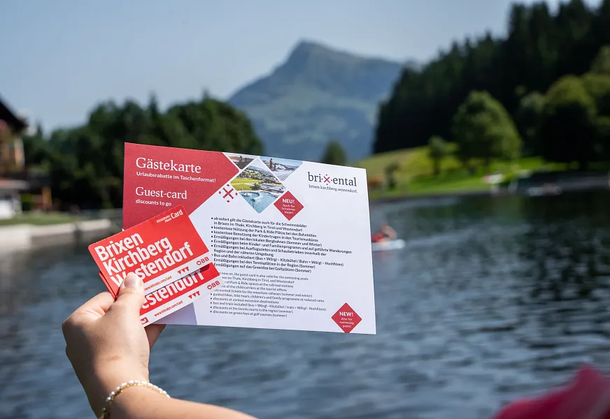 Person holds a guest card by a lake with mountains in the background in Tyrol, Kitzbühel.