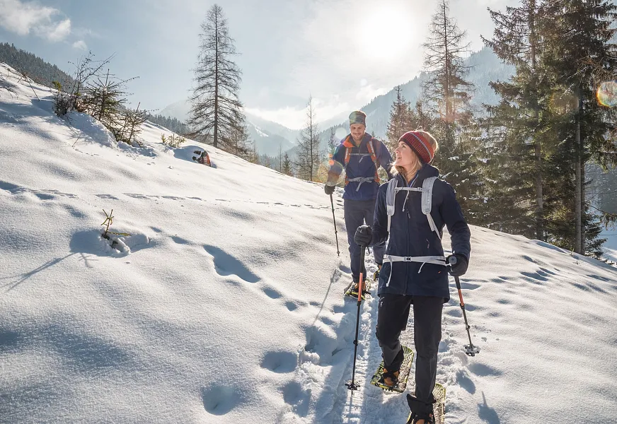 Zwei Menschen wandern im Schnee mit Skistöcken durch eine verschneite Berglandschaft unter klarem Himmel.