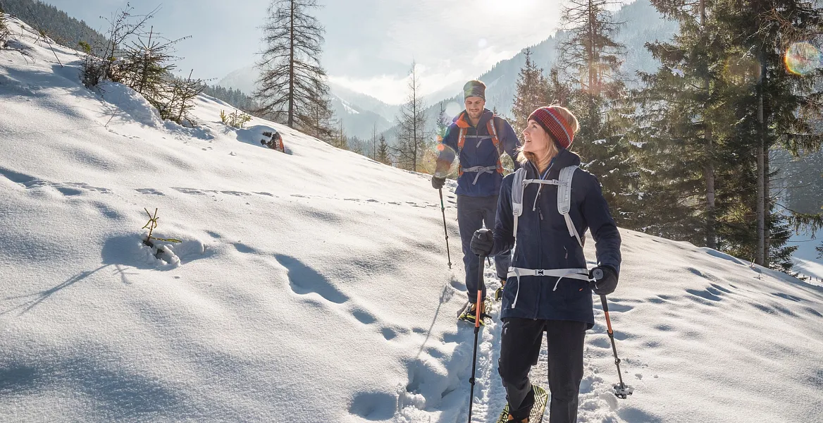 Zwei Menschen wandern im Schnee mit Skistöcken durch eine verschneite Berglandschaft unter klarem Himmel.