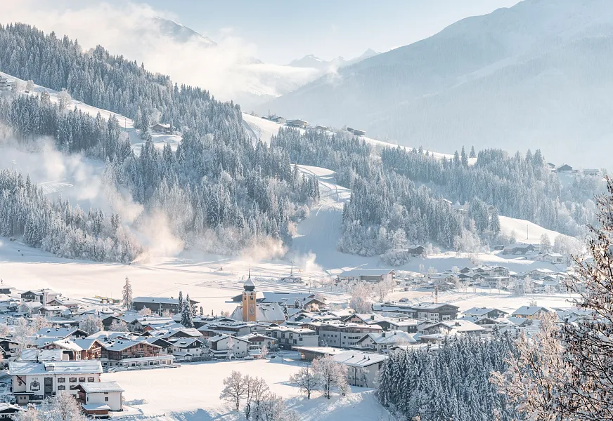 Verschneite Alpenlandschaft mit Dorf und Kirche in Kitzbühel, Tirol, umgeben von Bergen unter klarem Himmel.