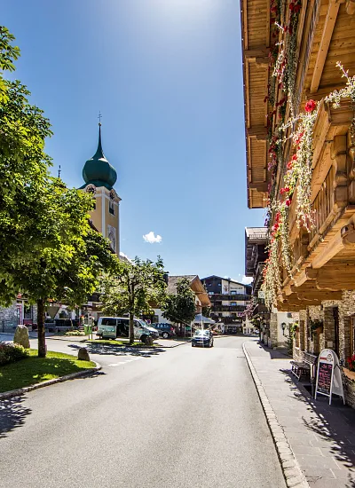 Straße in Kitzbühel mit traditionellen Gebäuden, Blumen und Kirchturmsicht an einem sonnigen Tag.