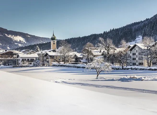Verschneite Berglandschaft mit Dorfkirche in den Alpen, umgeben von Wäldern und Hängen, unter klarem Himmel.